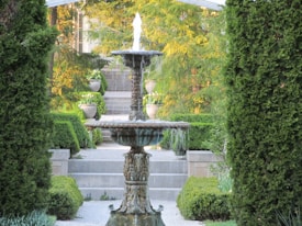 An ornate, tiered garden fountain surrounded by lush greenery and tall hedges. The fountain is elegantly crafted, with water gently cascading down multiple levels. In the background, steps lead up to what appears to be a garden or park area, flanked by stone planters filled with foliage.