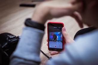 A person is holding a smartphone with a red case, recording a video. The screen displays a scene being filmed, showing a man behind a table with various equipment. Another object, possibly a camera, is partially visible on the table next to the person holding the phone.
