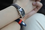 A close-up view of a wrist with two watches. One watch features a leather strap with a rectangular black dial showcasing metallic hour markers and hands. The other watch has a rubber strap, red and black in color, with a round emblem or button. The background is a soft, neutral shade, possibly clothing or a surface.