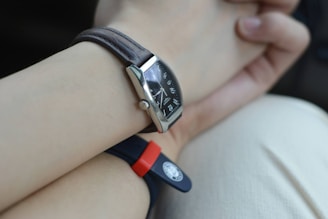A close-up view of a wrist with two watches. One watch features a leather strap with a rectangular black dial showcasing metallic hour markers and hands. The other watch has a rubber strap, red and black in color, with a round emblem or button. The background is a soft, neutral shade, possibly clothing or a surface.