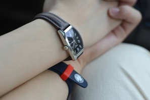 A close-up view of a wrist with two watches. One watch features a leather strap with a rectangular black dial showcasing metallic hour markers and hands. The other watch has a rubber strap, red and black in color, with a round emblem or button. The background is a soft, neutral shade, possibly clothing or a surface.