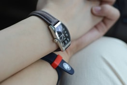 A close-up view of a wrist with two watches. One watch features a leather strap with a rectangular black dial showcasing metallic hour markers and hands. The other watch has a rubber strap, red and black in color, with a round emblem or button. The background is a soft, neutral shade, possibly clothing or a surface.