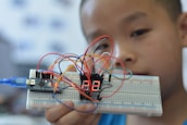 A young person holding up a breadboard with an electronic circuit. The setup includes a microcontroller connected to a digital display, and several colorful wires are attached to various components.