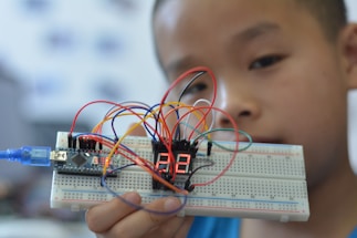 A young person holding up a breadboard with an electronic circuit. The setup includes a microcontroller connected to a digital display, and several colorful wires are attached to various components.