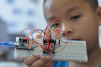 A young person holding up a breadboard with an electronic circuit. The setup includes a microcontroller connected to a digital display, and several colorful wires are attached to various components.