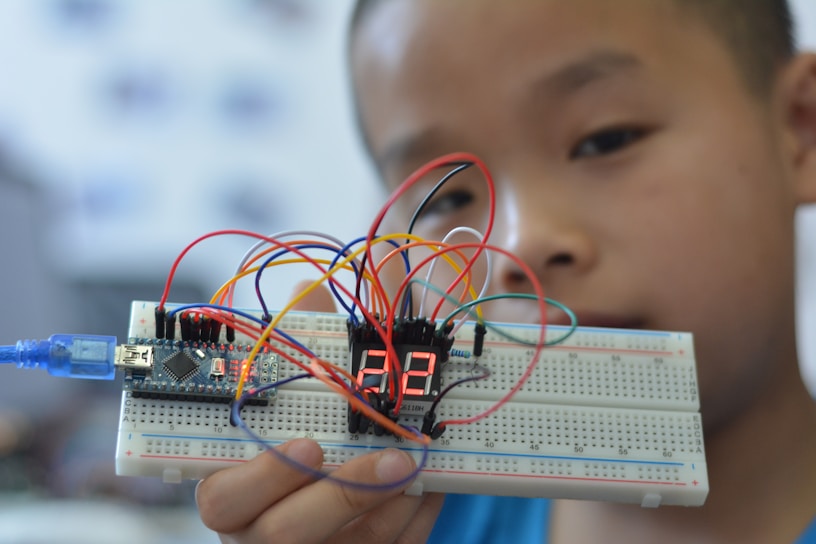 A young person holding up a breadboard with an electronic circuit. The setup includes a microcontroller connected to a digital display, and several colorful wires are attached to various components.