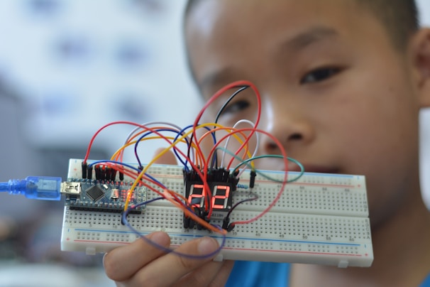 A young person holding up a breadboard with an electronic circuit. The setup includes a microcontroller connected to a digital display, and several colorful wires are attached to various components.