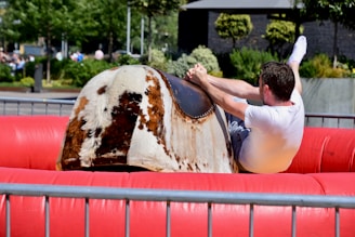 A man is riding a mechanical bull in an outdoor setting, surrounded by an inflatable red cushion. The scene is lively with green trees and other people in the background.