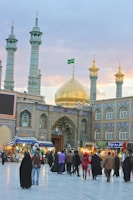Several people are walking and standing in front of a grand mosque with intricate tile work and multiple minarets. The mosque has a prominent golden dome with a green flag atop. There are also several shops and stalls on either side of the entrance.