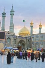 Several people are walking and standing in front of a grand mosque with intricate tile work and multiple minarets. The mosque has a prominent golden dome with a green flag atop. There are also several shops and stalls on either side of the entrance.