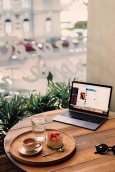 A cozy cafe scene with a wooden table holding a tray carrying a cup of coffee, a glass of water, and a jar of dessert topped with strawberries. Next to the tray is an open laptop showing a social media or entertainment screen. Sunglasses rest nearby, with green plants and a blurred street view visible through the window, creating a relaxed and inviting atmosphere.