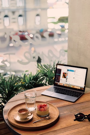 A cozy cafe scene with a wooden table holding a tray carrying a cup of coffee, a glass of water, and a jar of dessert topped with strawberries. Next to the tray is an open laptop showing a social media or entertainment screen. Sunglasses rest nearby, with green plants and a blurred street view visible through the window, creating a relaxed and inviting atmosphere.