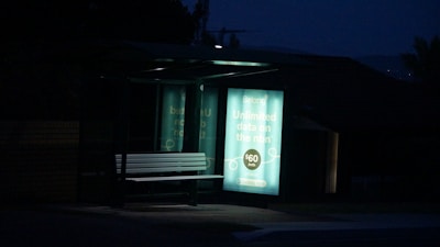 A dimly lit bus stop bench is illuminated by an advertisement panel displaying an internet service promotion. The surrounding area is dark, with the advertisement text prominently visible.