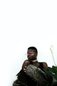 A person is wearing stylish sunglasses and large earrings, with a fur coat draped over their shoulders. The background is minimalistic and bright, with a large green plant visible to the right.