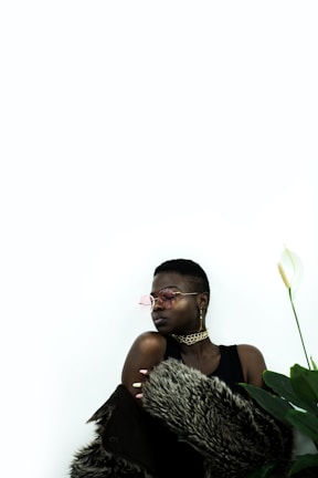 A person is wearing stylish sunglasses and large earrings, with a fur coat draped over their shoulders. The background is minimalistic and bright, with a large green plant visible to the right.