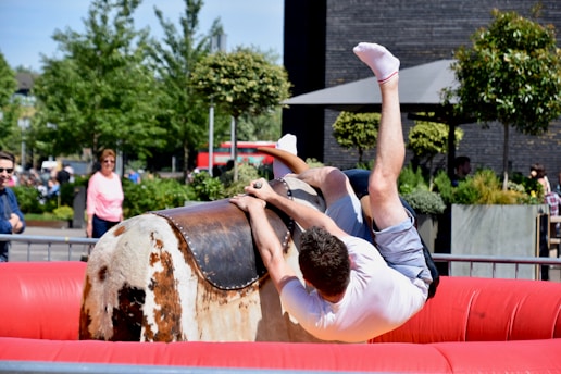 A person is falling off a mechanical bull in an outdoor setting. The bull is situated in a red inflatable ring, and the scene is surrounded by people watching. There are trees and plants in the background, along with a modern building.