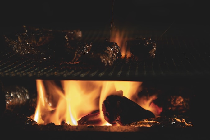 Dark, grilled meat pieces are positioned on a grate above a lively orange and yellow fire, creating a warm and smoky setting.