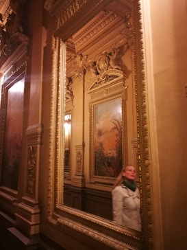 A richly decorated interior featuring ornate gold-framed mirrors, intricate moldings, and classic wall art with a natural theme. A person in a white coat walks past the mirrors, adding a modern element to the historic setting.