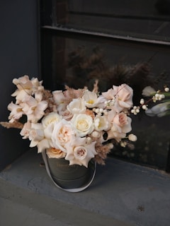 A bouquet of soft pink, cream, and white roses with dried floral accents is arranged in a dark round hatbox, placed on a windowsill. The background is a muted gray wall and window, lending an elegant and understated setting to the delicate arrangement.