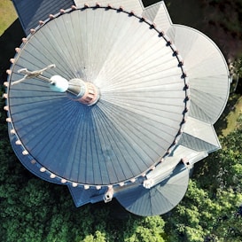 An aerial view of a large, circular, steel roof with a prominent spire in the center. The architectural structure is surrounded by trees, and the roof features radial patterns with visible segments and an intricate finial at the top.