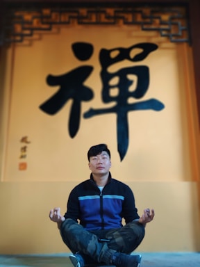 A person is sitting cross-legged in a meditative pose in front of a wall with large Chinese characters. The individual is wearing a dark jacket with a blue stripe and camo pants. The wall is painted in warm tones with a decorative pattern near the top.