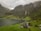 A scenic landscape features a small village nestled between lush green fields and a fjord. Towering mountains, partially covered in mist, surround the area with cascading waterfalls running down their sides. The village includes a few quaint houses and farm buildings scattered across the landscape.