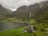 A scenic landscape features a small village nestled between lush green fields and a fjord. Towering mountains, partially covered in mist, surround the area with cascading waterfalls running down their sides. The village includes a few quaint houses and farm buildings scattered across the landscape.