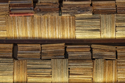 A collection of vintage books and magazines stacked on shelves. The covers and spines exhibit significant wear and aging, with faded colors and visible text. Some books are stacked horizontally while others are vertically arranged, creating a textured and layered appearance.