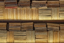 A collection of vintage books and magazines stacked on shelves. The covers and spines exhibit significant wear and aging, with faded colors and visible text. Some books are stacked horizontally while others are vertically arranged, creating a textured and layered appearance.