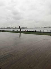 A wet wooden deck extends towards a waterfront under a cloudy, overcast sky. In the distance, industrial structures and cranes can be seen across the water.
