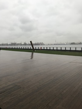 A wet wooden deck extends towards a waterfront under a cloudy, overcast sky. In the distance, industrial structures and cranes can be seen across the water.