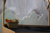 A woven basket filled with orange and green woolen yarn sits on a wooden structure. The background features a soft, blurred depiction of mountain-like shapes, possibly part of a fabric backdrop.