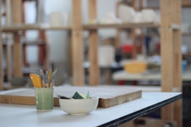 A focused view of an art studio workspace with a variety of sculpting tools in a green cup, a white bowl containing wet sponges, and a wooden tray on a white table. The background features wooden shelves filled with various art supplies, creating a busy yet organized atmosphere.