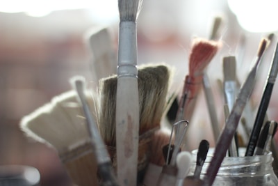 A close-up view of various paintbrushes, with soft focus creating a dreamy, artistic atmosphere. The brushes are gathered in a container, with bristles showing signs of use. The lighting is gentle, enhancing the textures and colors of the brushes.