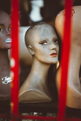 Several mannequin heads are arranged close to each other, featuring painted facial features like eyes and lips. The heads show signs of wear and are surrounded by a red frame or structure.