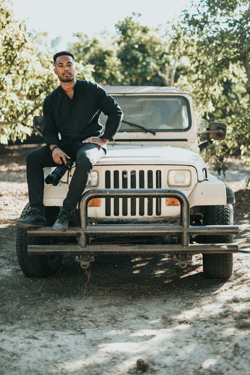 man sitting on white Jeep Wrangler SUV near trees during daytime