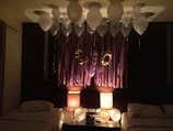 A room is decorated with white balloons attached to the ceiling and purple streamers hanging from the wall. Gold foil balloons form the numbers '20'. Two soft-lit lamps on each side of a small table cast a warm glow over the scene. The room includes two beds, each with white linens, suggesting a celebratory or festive atmosphere.