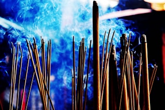 A cluster of incense sticks releasing swirls of blue smoke, creating an ethereal atmosphere. The background features a blurred and indistinct setting, enhancing the contrast between the incense and the vibrant smoke.