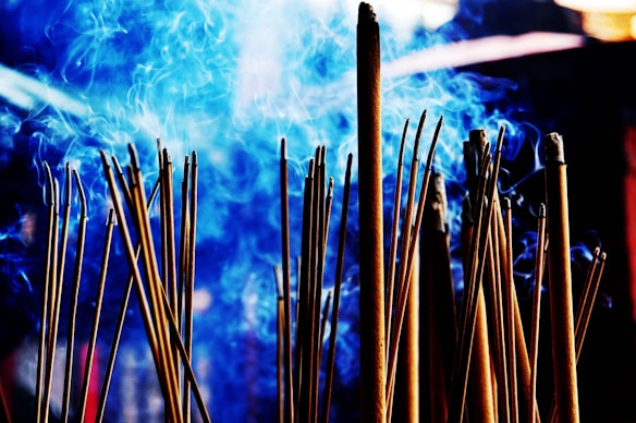 A cluster of incense sticks releasing swirls of blue smoke, creating an ethereal atmosphere. The background features a blurred and indistinct setting, enhancing the contrast between the incense and the vibrant smoke.