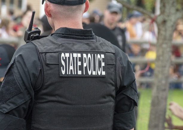 A state police officer is wearing a black uniform with a radio visible on the shoulder, standing outdoors with a crowd in the background. The focus is on the back of the officer, highlighting the words 'State Police' on the vest.