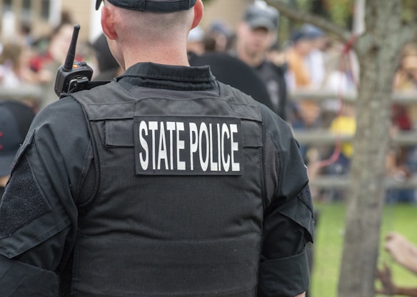 A state police officer is wearing a black uniform with a radio visible on the shoulder, standing outdoors with a crowd in the background. The focus is on the back of the officer, highlighting the words 'State Police' on the vest.