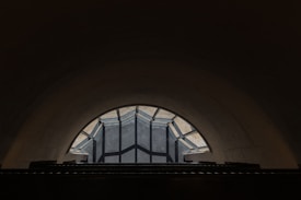 A dark underground tunnel with a large, arched ceiling made of concrete. At one end of the tunnel, a geometric glass structure allows a limited amount of natural light to enter, creating an intriguing pattern of shadows. The contrast between the dark interior and the light from the glass is visually striking.