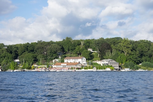 A serene lakeside scene features a cluster of buildings along the water's edge, backed by lush green trees. The landscape includes a mix of residential homes and smaller structures, with calm water in the foreground. The sky is partially cloudy, adding to the tranquil atmosphere.