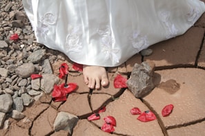 A bare foot partially covered by a white dress embroidered with floral designs is stepping on cracked, dry earth. Surrounding the foot are scattered red flower petals and rocks, creating a contrast between the delicate petals and the harsh terrain.