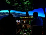 The image captures the interior of an airplane cockpit at night. It includes illuminated control panels and screens, displaying various flight instruments with a view of a landscape outside that suggests flight simulation.