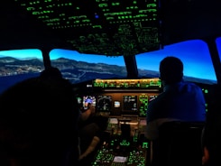 The image captures the interior of an airplane cockpit at night. It includes illuminated control panels and screens, displaying various flight instruments with a view of a landscape outside that suggests flight simulation.