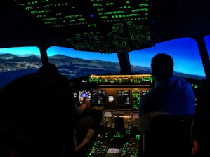 The image captures the interior of an airplane cockpit at night. It includes illuminated control panels and screens, displaying various flight instruments with a view of a landscape outside that suggests flight simulation.