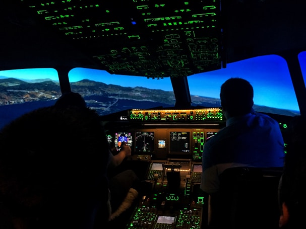 The image captures the interior of an airplane cockpit at night. It includes illuminated control panels and screens, displaying various flight instruments with a view of a landscape outside that suggests flight simulation.