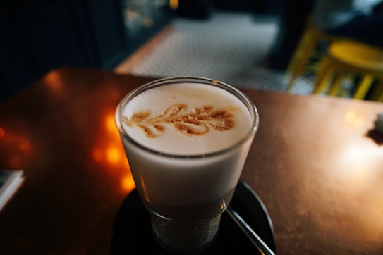 A glass of latte topped with a leaf-shaped latte art design is placed on a saucer with a spoon. The background features a blurred interior with warm lighting and tiled flooring.