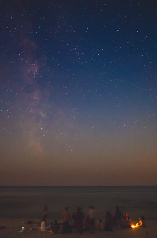 A group of friends gathered around a campfire on the beach under a star-filled sky.